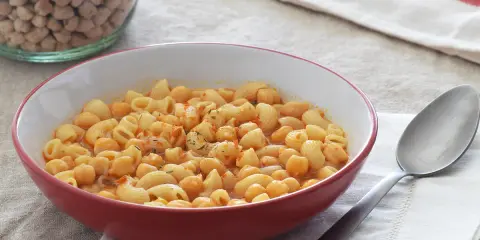 Un plato humeante de pasta e ceci, con garbanzos y macarrones en una salsa rojiza, junto a un tarro de garbanzos secos.